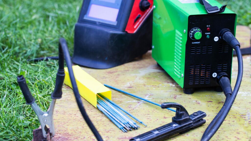 Essential welding safety equipment including welding helmet, electrodes, and green welding machine on grass
